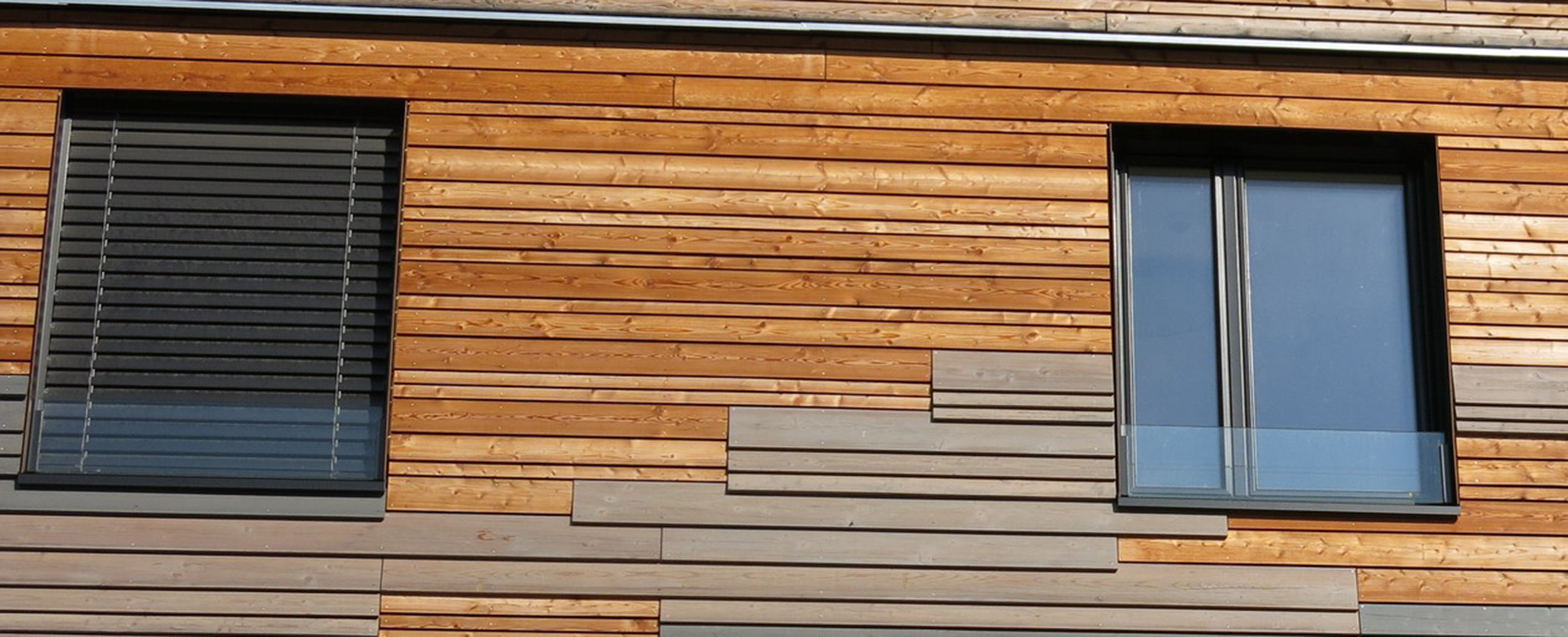 Wooden façade made of different types of wood with two windows, one of which has sun protection.