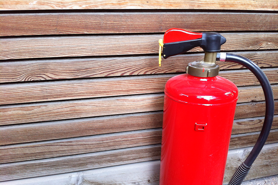 Ein knallroter Feuerlöscher steht vor einer Holzfassade.